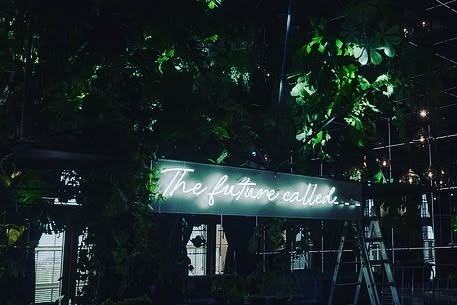 Neon sign reading 'The future called' installed among trees at night for Pinterest
